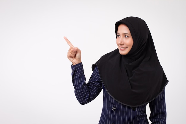 Mujer de negocios musulmana acertada sonriente feliz confiada que destaca; retrato de estudio de dedo acusador de mujer de negocios islámica; mujer de negocios asiática, persona de negocios femenina apunta al espacio