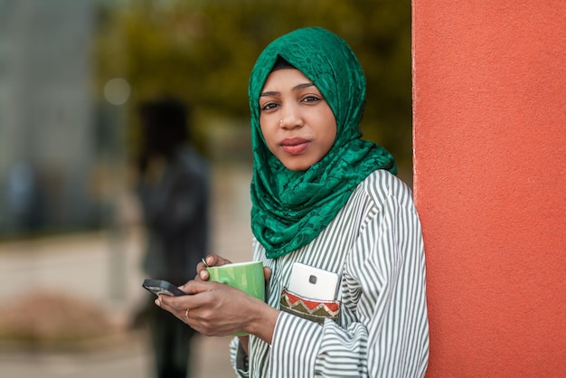 Mujer de negocios musulmana africana con hiyab verde usando un teléfono inteligente durante un descanso para tomar café del trabajo afuera. foto de alta calidad