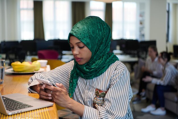 mujer de negocios musulmana africana que usa un hiyab verde usando un teléfono móvil mientras trabaja en una computadora portátil en el área de relajación en la moderna oficina de inicio de planta abierta. Diversidad, concepto multirracial