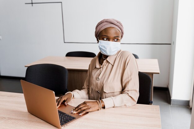 Mujer de negocios musulmana negra en máscara médica con ordenador portátil.