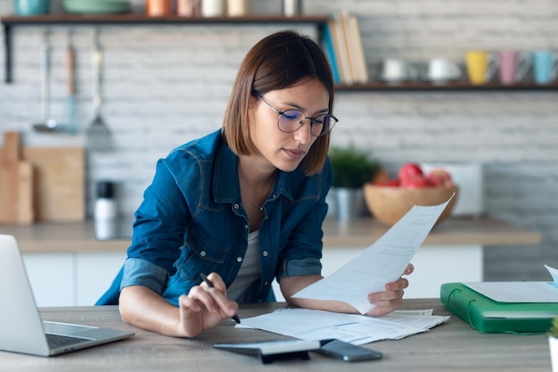 Mujer de negocios muy joven que trabaja con una computadora mientras consulta algunas facturas y documentos en la cocina de casa