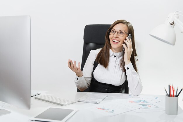 Mujer de negocios muy sonriente en traje sentada en el escritorio, trabajando en una computadora contemporánea con documento en la oficina ligera, hablando por teléfono móvil, manteniendo una conversación agradable