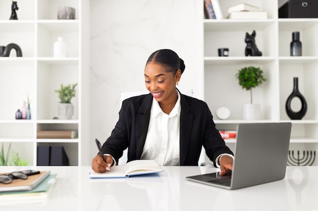 Mujer de negocios negra alegre o psicóloga trabajando en una computadora portátil y tomando notas durante el trabajo en línea