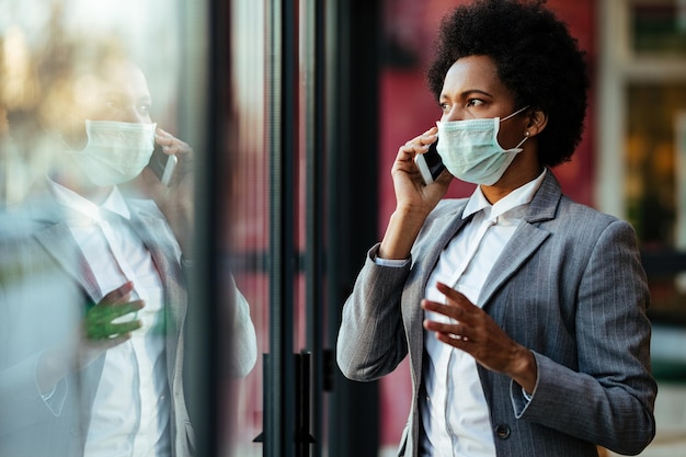 Mujer de negocios negra con máscara protectora en la cara y hablando por teléfono inteligente mientras mira por la ventana