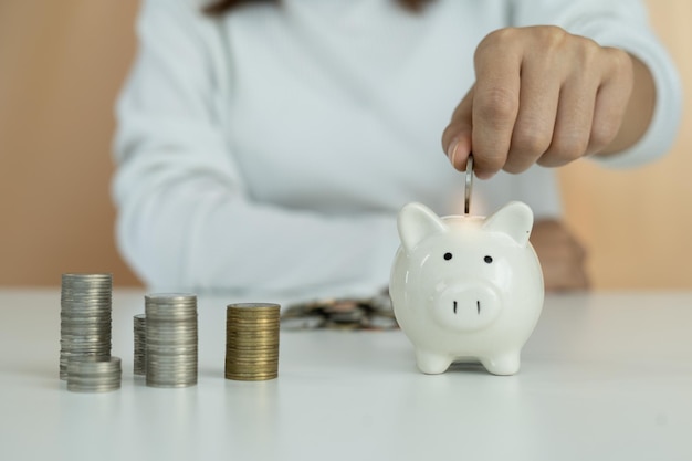 La mujer de negocios o el ama de llaves está poniendo monedas en una alcancía con monedas apiladas en la mesa mujer para recolectar ahorros como capital comercial o ahorrar dinero para el futuro Concepto de ahorro de dinero