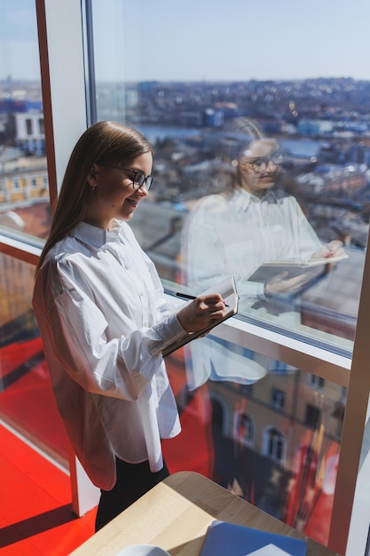 Mujer de negocios pensativa con bloc de notas vestido con ropa formal de pie cerca de la ventana panorámica en una empresa corporativa y pensando en la inversión comercial mujer de negocios pensativa con gafas