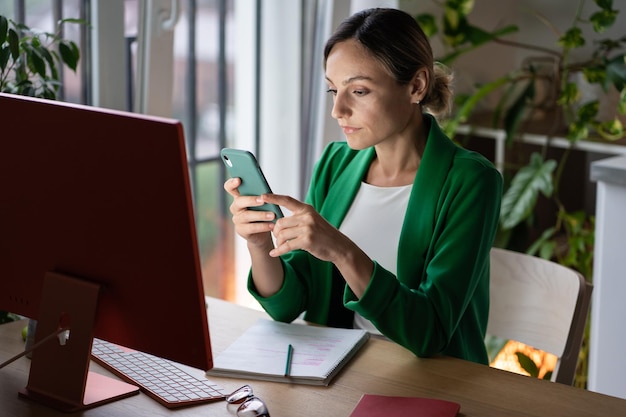 Mujer de negocios pensativa sentada en la mesa frente a la computadora usa el teléfono inteligente para noticias de desplazamientos de trabajo