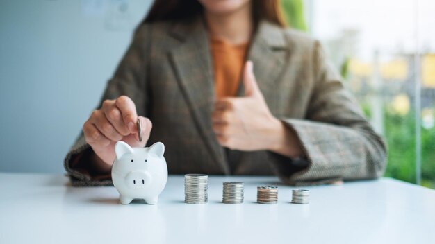 Una mujer de negocios poniendo monedas en la alcancía mientras muestra los pulgares para arriba signo de mano con pila de monedas sobre la mesa para ahorrar dinero y concepto financiero