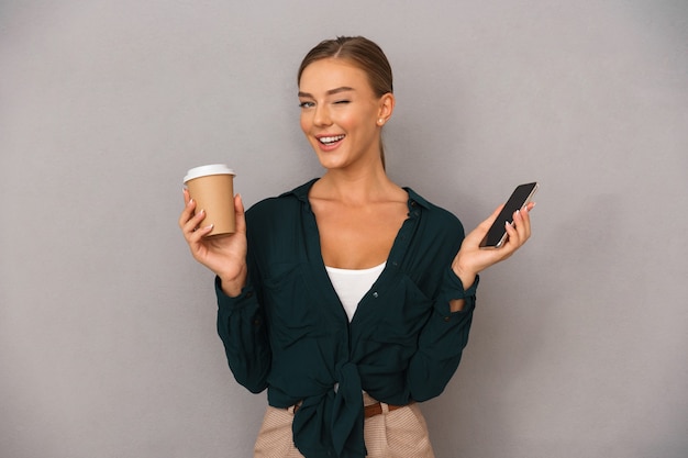 Mujer de negocios posando aislada sobre fondo de pared gris tomando café con teléfono móvil.