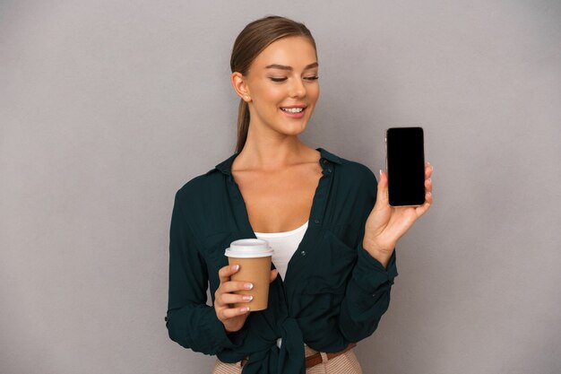 Mujer de negocios posando aislado sobre fondo de pared gris tomando café mostrando la pantalla del teléfono móvil.