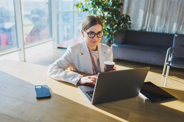 Una mujer de negocios positiva con una chaqueta está sentada en una mesa trabajando en una computadora portátil en una oficina luminosa Una joven europea está sentada en una mesa Trabajo independiente y remoto Estilo de vida moderno de la mujer