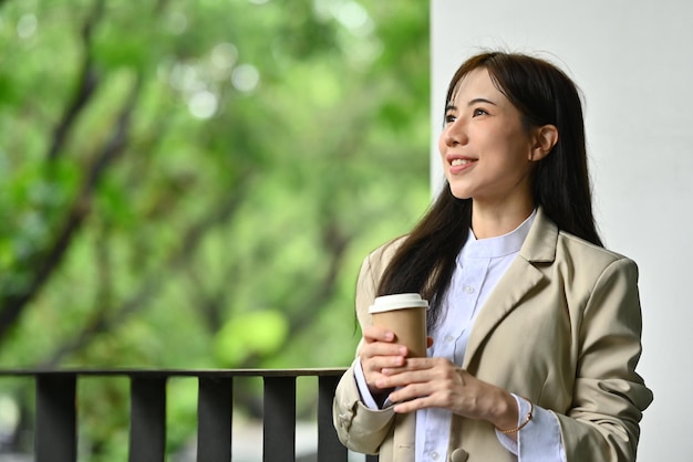 Mujer de negocios positiva con una taza de papel de pie fuera del edificio de oficinas y mirando lejos pensando