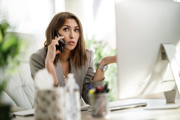 Una mujer de negocios preocupada hablando por teléfono inteligente mientras trabaja en la computadora en su oficina en casa.