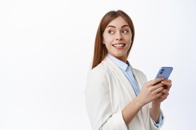 Mujer de negocios profesional sostiene el teléfono inteligente y gira la cabeza a un lado en el texto promocional, mira copyspace con una sonrisa de satisfacción, de pie con el teléfono móvil sobre fondo blanco