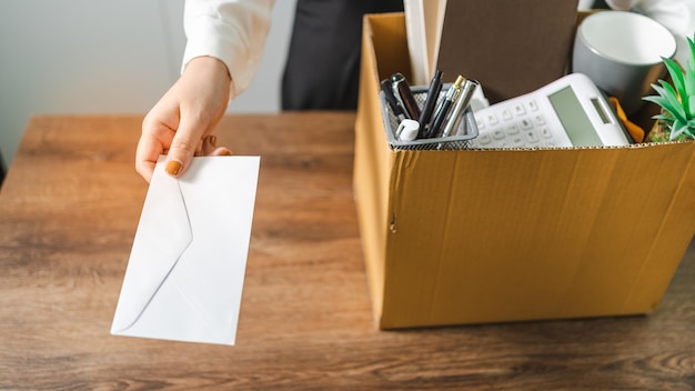 Mujer de negocios que envía una carta de renuncia y empaca cosas Renuncia Deprimir o llevar una caja de cartón comercial por escritorio en la oficina Cambio de trabajo o despedida de la empresa