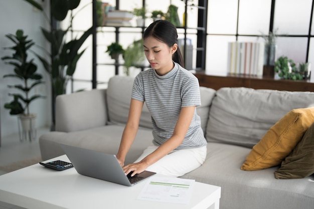 Mujer de negocios que trabaja desde casa, trabaja en línea en una computadora portátil, mujer de negocios asiática que trabaja en un sofá, negocios en línea con distanciamiento social, reunión en línea con computadora portable.