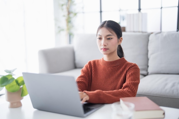 Mujer de negocios que trabaja desde casa, trabaja en línea en una computadora portátil, mujer de negocios asiática que trabaja en un sofá, negocios en línea con distanciamiento social, reunión en línea con computadora portable.