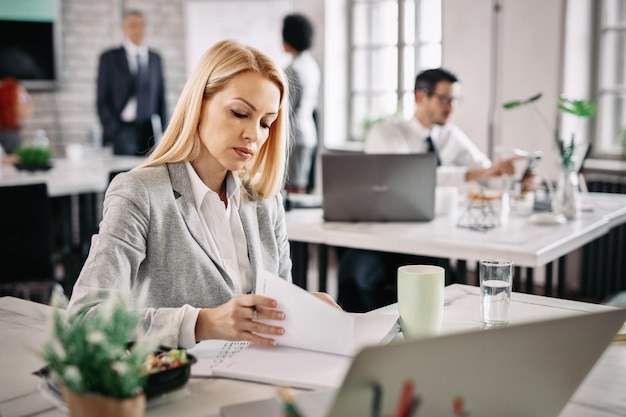 Mujer de negocios que trabaja en el escritorio de la oficina y se prepara para la próxima reunión mientras revisa sus notas Hay personas en el fondo