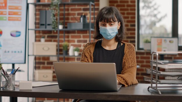 Mujer de negocios que trabaja con portátiles y gráficos para planificar el proyecto y la presentación. Emprendedor con mascarilla usando tecnología y documentos sobre la mesa para diseñar la estrategia de marketing.