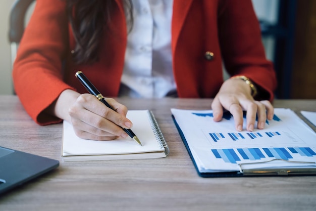 Mujer de negocios que usa una computadora portátil para trabajar y escribe en un cuaderno con bolígrafo en la oficina con el móvil junto a los papeles