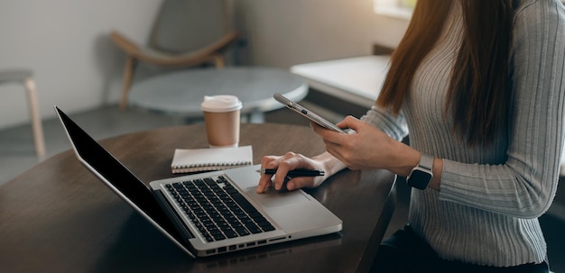 Mujer de negocios que usa un teléfono inteligente y una computadora portátil para trabajar y escribir en un cuaderno con un bolígrafo en un café con una taza de café al lado de la computadora portátil