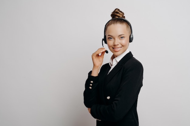 Mujer de negocios relajada en traje oscuro con auriculares inalámbricos oscuros durante la conversación web con colegas, posando con expresión facial positiva en estudio sobre fondo gris