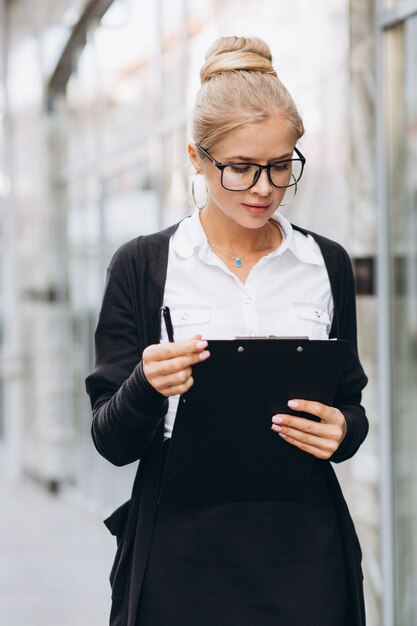 Mujer de negocios rubia de moda hermosa en vidrios que habla por el teléfono móvil y la situación fuera de un edificio de oficinas.