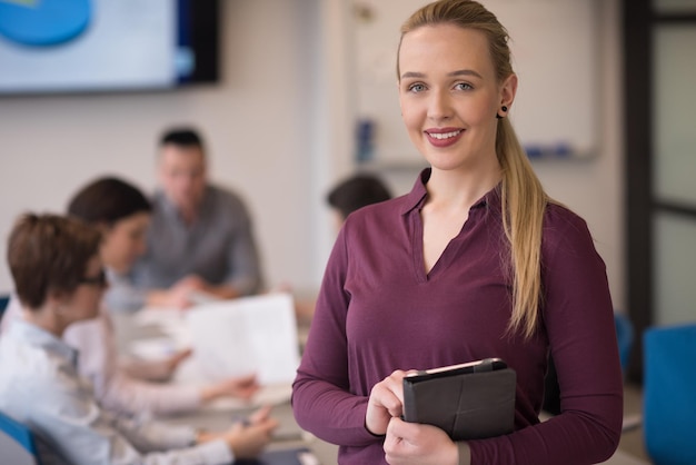 Mujer de negocios rubia con ropa informal trabajando en una tableta en el moderno interior de la oficina de negocios de inicio. Grupo de jóvenes en reunión de equipo borrosa en segundo plano.