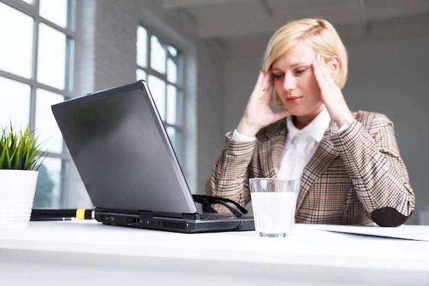 Una mujer de negocios rubia vestida con un traje elegante tiene dolor de cabeza trabajando usando una pastilla efervescente en el agua en un lugar de trabajo de vidrio con una computadora portátil en una oficina blanca