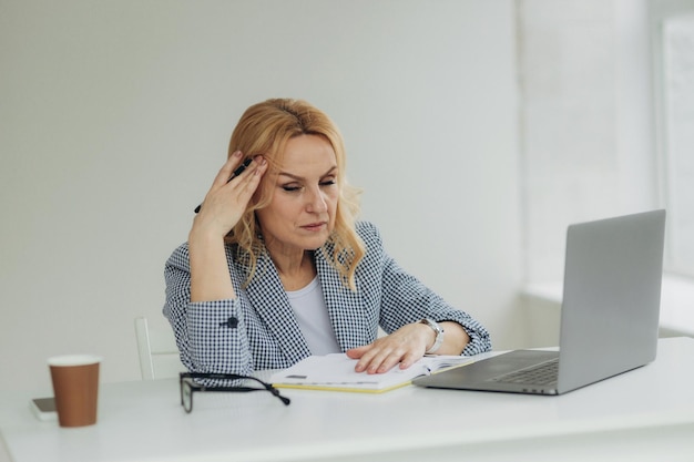 Mujer de negocios senior cansada está usando una computadora portátil para trabajar en interiores Una hermosa señora mayor tiene dolor de cabeza por el exceso de trabajo Mujer madura de 50 años