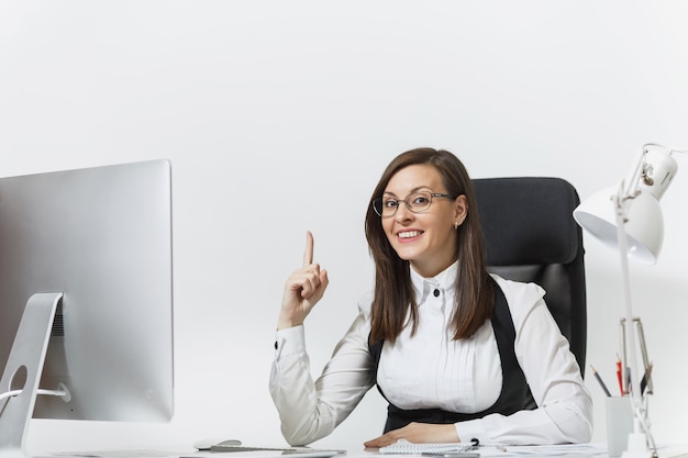 Mujer de negocios sentada en el escritorio, trabajando en una computadora moderna con documentos en la oficina de luz, apuntando hacia arriba con el dedo índice