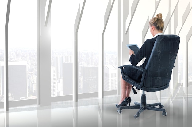 Mujer de negocios sentada en una silla giratoria con tableta contra la habitación con una gran ventana mirando a la ciudad