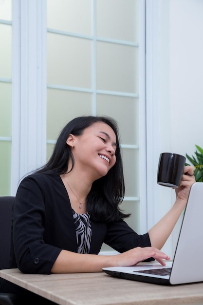 Mujer de negocios sentada en una silla mientras sonríe dan sosteniendo un vaso frente a la computadora portátil