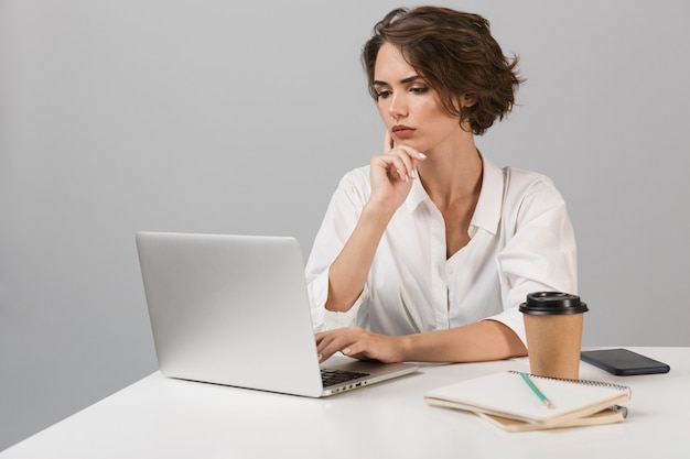 Mujer de negocios seria posando aislada sobre pared gris sentada en la mesa usando laptop