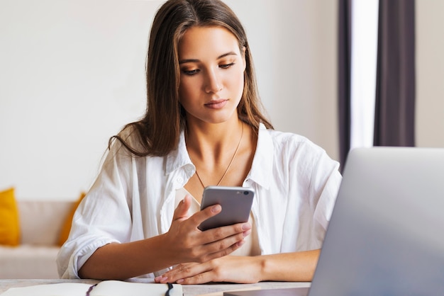 Mujer de negocios se sienta a la mesa frente a la computadora portátil, escribe un mensaje con un teléfono inteligente, realiza correspondencia comercial.