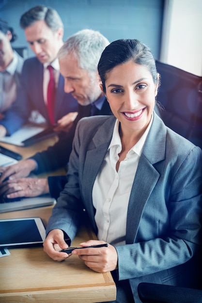 Mujer de negocios sonriendo a la cámara y empresarios interactuando en segundo plano en la sala de conferencias durante la reunión