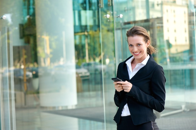 Mujer de negocios sonriendo con teléfono celular fuera del edificio de oficinas