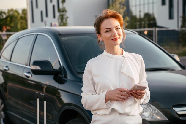 Mujer de negocios sonriente con bolsa de viaje sosteniendo un teléfono móvil cerca del auto. viajes de negocios