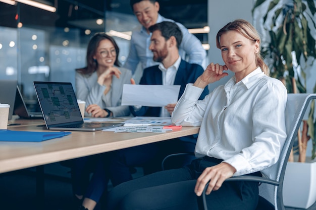 Una mujer de negocios sonriente y confiada sentada en una reunión en la oficina con sus colegas en el fondo