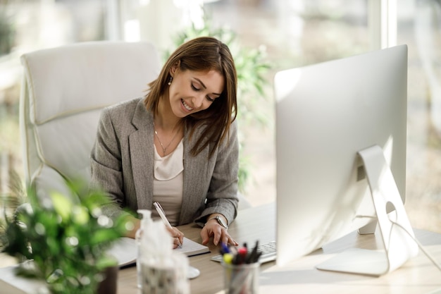 Una mujer de negocios sonriente escribe algo en un cuaderno mientras usa una computadora en la oficina de su casa.