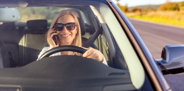 Mujer de negocios sonriente con gafas de sol hablando por teléfono móvil mientras conduce un automóvil moderno Situación peligrosa en la carretera Se necesita concentración