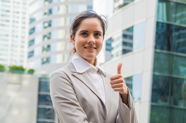 Foto mujer de negocios sonriente gestando los pulgares hacia arriba mientras está de pie contra el edificio