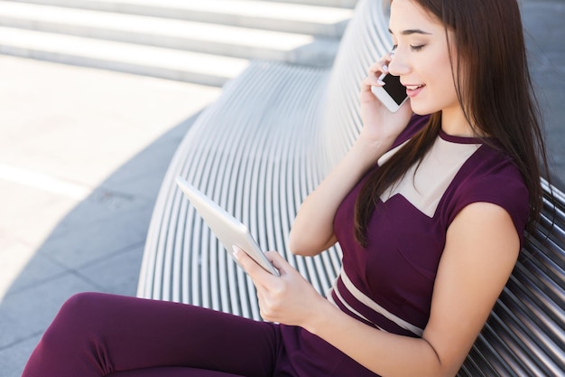 Mujer de negocios sonriente hablando por teléfono y trabajando en una tableta, sentada en un banco al aire libre cerca del moderno centro de oficinas, copiando espacio. Trabajo independiente, concepto de estilo de vida de niña ocupada joven, espacio de copia