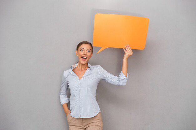 Mujer de negocios sonriente hermosa que se coloca sobre fondo gris de la pared que sostiene la burbuja del discurso.