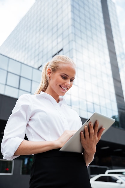 Mujer de negocios sonriente joven que usa la tableta que se coloca en el edificio de oficinas al aire libre