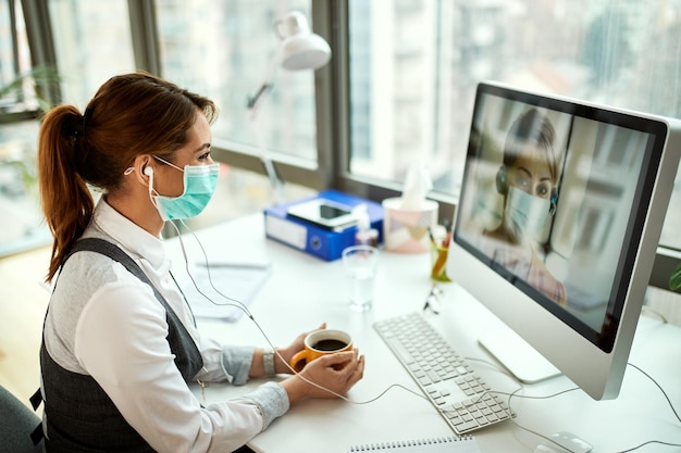 Mujer de negocios sonriente con mascarilla usando computadora y haciendo videollamadas mientras toma un descanso para tomar café en la oficina