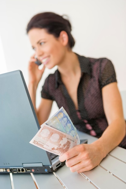 Foto mujer de negocios sonriente con papel moneda usando teléfono por portátil en el escritorio