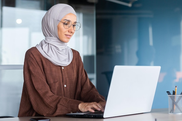 Mujer de negocios sonriente y soñadora que trabaja dentro de la oficina con una mujer portátil en la oficina de hiyab y anteojos