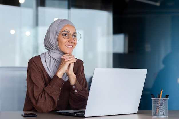 Mujer de negocios sonriente y soñadora que trabaja dentro de la oficina con una mujer portátil en la oficina de hiyab y anteojos