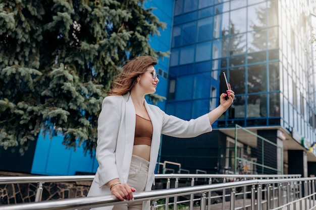 Mujer de negocios sonriente con traje blanco y gafas de sol haciendo videollamadas a un amigo usando el teléfono durante el descanso de pie cerca del edificio de oficinas moderno